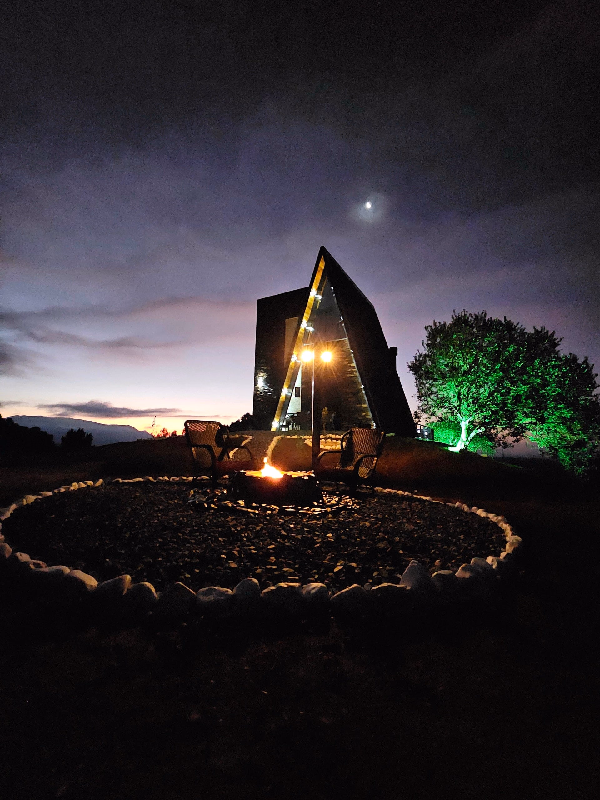 Chalé A-frame iluminado ao anoitecer, com fogo de chão aceso em círculo de pedras e iluminação verde na árvore ao lado