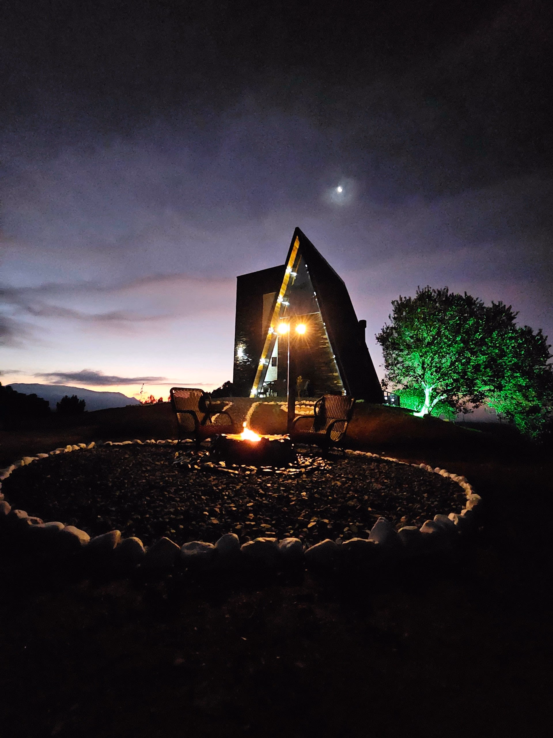 Chalé A-frame iluminado ao anoitecer, com fogo de chão aceso em círculo de pedras e iluminação verde na árvore ao lado
